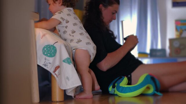 Toddler successfully climbing onto bed and reaching mattress edge while caregiver sits nearby holding toy showing progression and growing independence in home environment