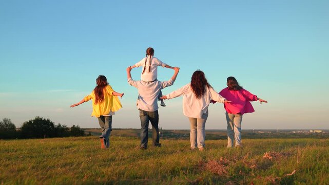 Family picnic nature, children parents running green grass towards sky. Parents, dad mom children play outdoors. Child on father's shoulders, mother children run together field. People walk on lawn