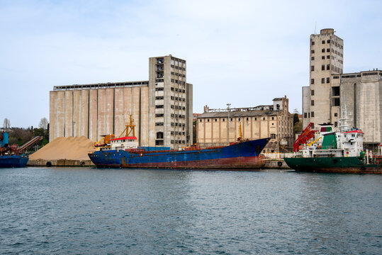 Cargo ships docked at a harbor near industrial buildings with storage silos on a clear day