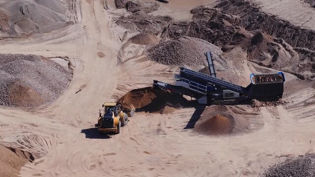 Aerial view of an arid quarry as a yellow wheel loader scoops and moves material beside a mobile screening plant. Conveyor forms conical stockpiles under midday sun.