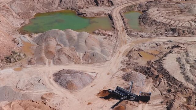 Aerial view of an open pit quarry shows terraced benching, dirt haul roads, greenish ponds, conical rock stockpiles, a mobile conveyor, a loader, and strong daylight shadows.