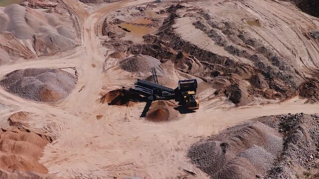 Aerial view of a quarry shows a mobile screener and loader moving. Conveyors sort stone into conical piles, graded stockpiles and benches ring the pit in daylight.