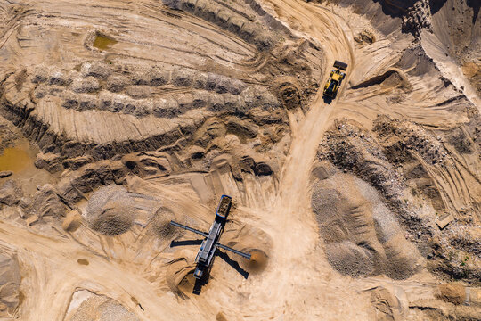 Aerial view of an open pit quarry shows a yellow haul truck climbing a dirt ramp. A mobile crusher with conveyors sits central amid gravel and sand stockpiles.