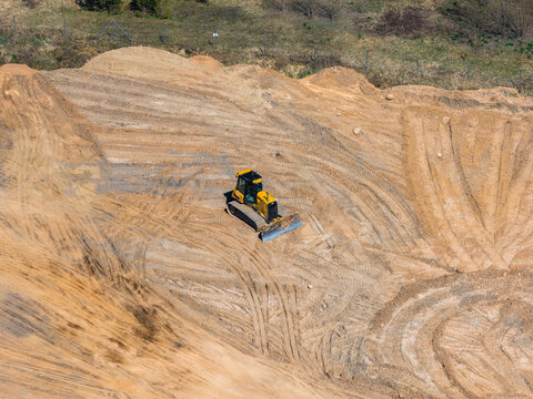 Aerial view shows a yellow and black bulldozer pushing a windrow across rippled, tire scored ground in a terraced open pit site under midday light in semi arid scrubland.