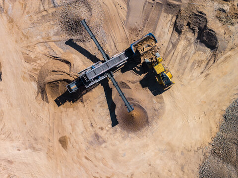 Aerial view of an open pit quarry shows a mobile crusher and cross conveyors. A yellow haul truck offloads into the hopper as belts form conical gravel mounds at midday.