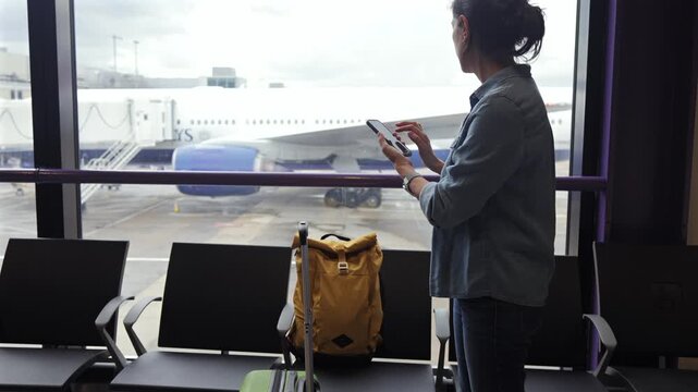 Woman passenger using a smartphone at the airport terminal while waiting for her flight, airplane in the background connected to the jet bridge at the boarding gate