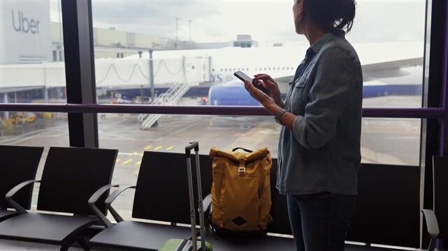Woman with luggage using her smartphone while looking out an airport window at a plane on the tarmac, waiting for departure in the boarding gate area before her flight