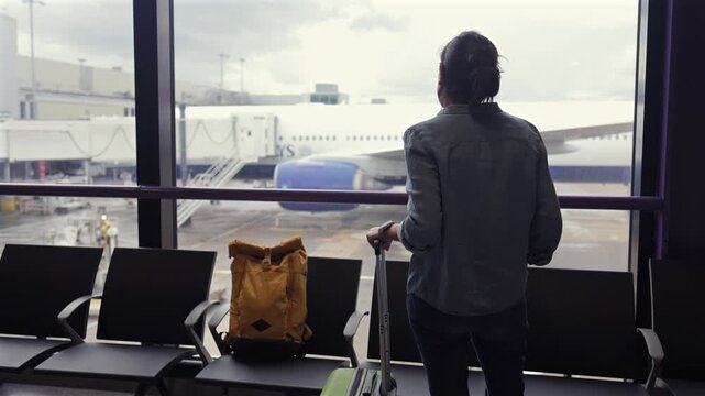Young woman passenger with luggage waiting for her flight at the airport terminal, looking through the window at the airplane ready for boarding at the departure gate