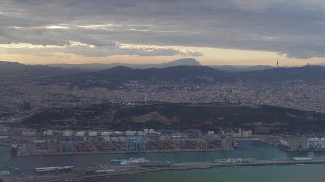 Barcelona skyline aerial view at sunset, capturing the urban landscape, montjuic hill, and the industrial harbor with cargo ships and shipping containers cranes