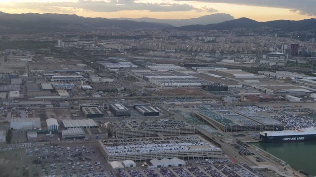 Aerial view of the barcelona industrial area with the port car terminal, freight shipping, and commercial vehicle depot against a stunning mountain sunset backdrop