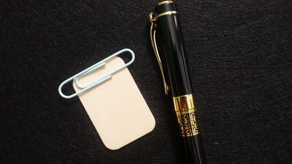 Fototapeta premium Black pen with gold accents rests near a paperclip holding a small note
