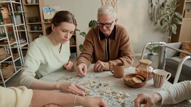 Arc handheld shot of young Caucasian caretaker assisting group of senior adults assembling jigsaw puzzle pieces on table in cozy care home room