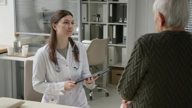 Medium shot of young adult female general practitioner filling out patients record on digital tablet listening to elderly man complaining about migraine in exam room