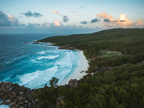 Aerial view of the pristine white sand beach of Grand Anse with turquoise waves crashing against granite rocks and lush tropical hills under a soft sunset sky in Grand Anse, La Digue, Seychelles.