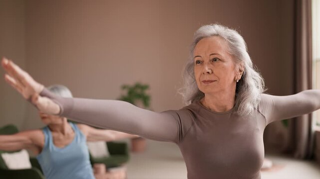 Focused women in their sixties doing yoga indoors, practicing warrior pose