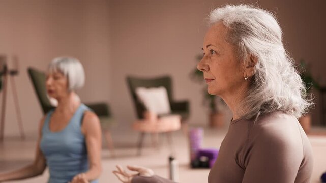 Focused senior women meditating together, sitting in lotus pose, yoga session