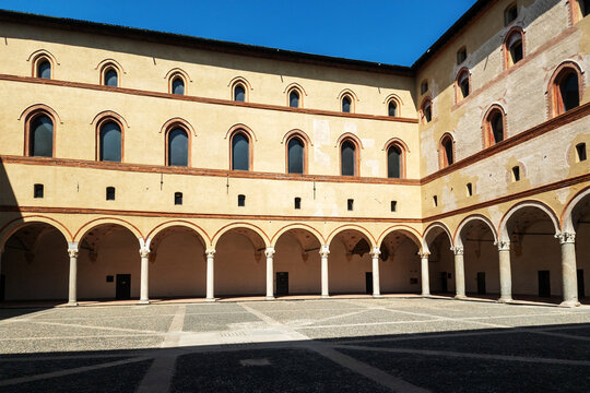 The historic courtyard of Castello Sforzesco in Milan features impressive Renaissance arcades with elegant columns and arched windows set against a vibrant blue sky
