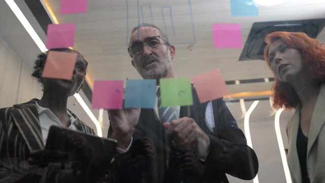 Multicultural business team brainstorming with sticky notes on glass wall, coworkers analyzing project strategy, creative coworkers collaborating in modern office, agile startup workflow concept.