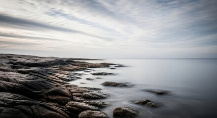 Fototapeta premium Rocks line the serene ocean shore under a cloudy sky at sunset