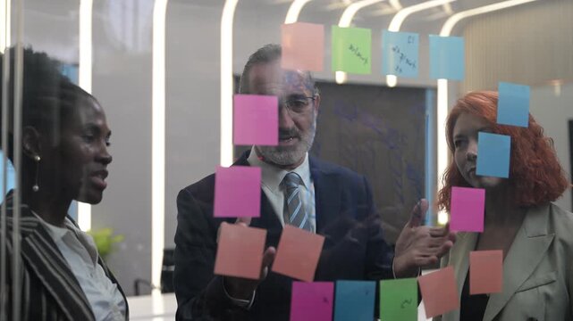 Multicultural business team brainstorming with sticky notes on glass wall, coworkers analyzing project strategy, creative coworkers collaborating in modern office, agile startup workflow concept.