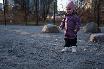 Fototapeta premium smiling toddler in a purple snowsuit and pink beanie stands on gravel at golden-hour in a quiet urban park playground, soft backlight, scattered boulders and bare trees evoke calm, chilly atmosphere
