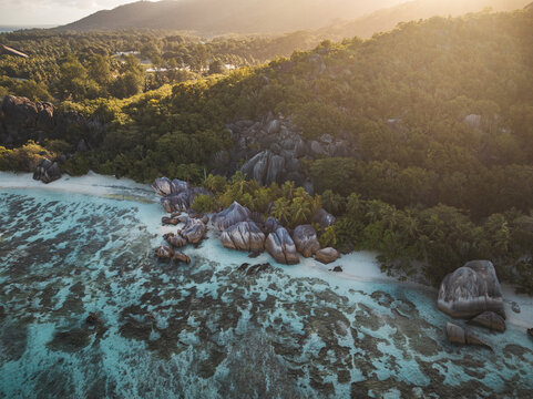 Aerial view of Anse Source d'Argent beach with its iconic granite boulders, turquoise coral reefs, and lush tropical forest under golden hour light at L'Union Estate, Seychelles.