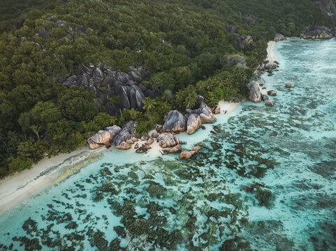 Aerial view of Anse Source d'Argent with its unique granite boulders, white sandy beach, and turquoise coral reef waters bordered by lush tropical forest in L'Union Estate, Seychelles.