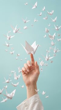 Human hand balancing a white origami paper crane on the index finger, surrounded by a swirling flock of flying small paper birds on a light blue background.
