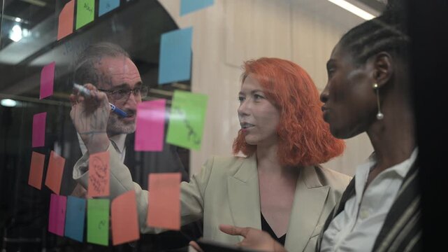 Multicultural business team brainstorming with sticky notes on glass wall, coworkers analyzing project strategy, creative coworkers collaborating in modern office, agile startup workflow concept.