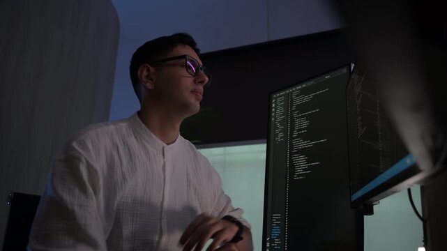 Focused young female software developer programming code on multiple monitors in a dark home office at night. Professional woman engineer working on cyber security and data science tech project.