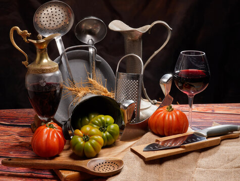 Still life, natura morta con pomodori su tavola imbandita con scenografia di vasi, pentole, utensili, calice di vino e brocca