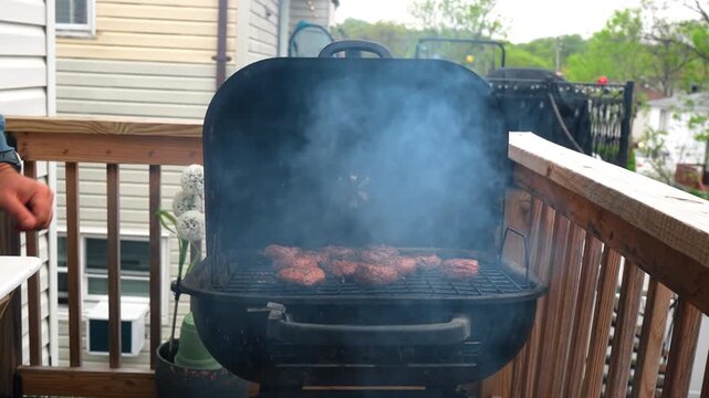 Barbecue grill smoking on a wooden deck at dusk. Wide shot of a black grill with smoke rising from meat patties on an outdoor balcony.