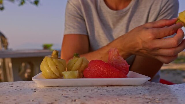 Close Up of Hand Taking Tropical Fruit from Plate with Pomelo and Pineapple at Seaside Table in Warm Light, Healthy Snack Lifestyle