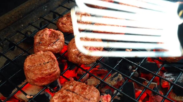 Flipping meat patties on charcoal grill with metal spatula. Action shot of metal spatula moving over meat patties grilling on hot charcoal with motion blur and glowing fire.