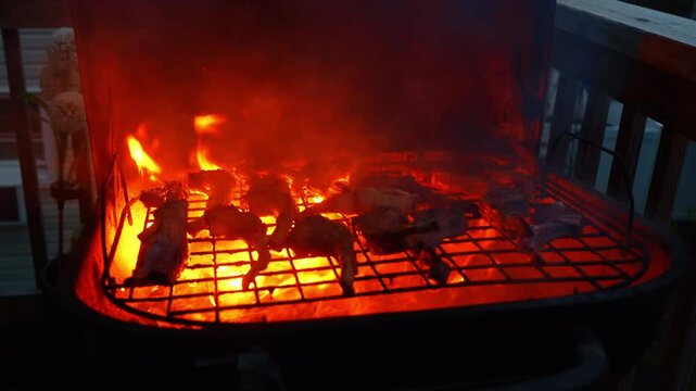 Meat chops cooking over intense glowing fire on grill. Red and orange flames engulfing meat pieces on a barbecue grate during evening.
