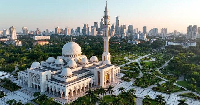 An aerial perspective reveals a stunning modern architectural structure with multiple white domes and a tall, elegant minaret, positioned within beautifully landscaped green gardens. Beyond this seren