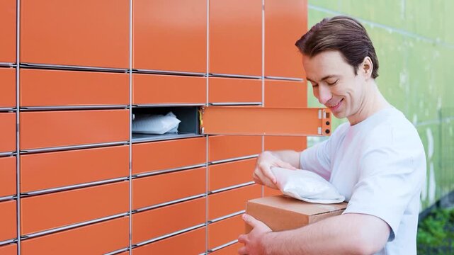 A smiling man is picking up packages from an orange automated parcel locker. He opens the compartment door and takes out the boxes, demonstrating easy self-service delivery. High quality 4k footage