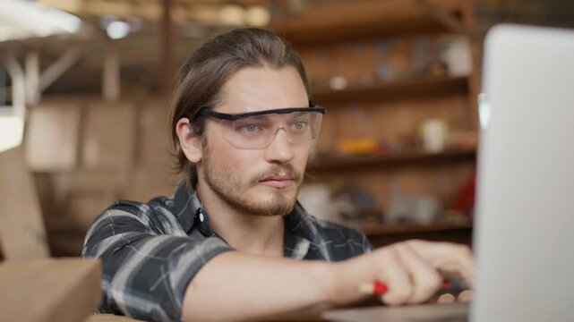 Professional male carpenter wearing safety goggles and using a laptop in a carpentry workshop. Skilled craftsman researching woodworking designs in a rustic industrial studio studio