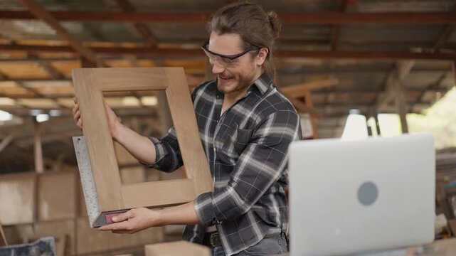 Professional carpenter using a square ruler to measure a wood frame during a video call on a laptop. Skilled craftsman presenting handmade project to client in a rustic industrial workshop studio
