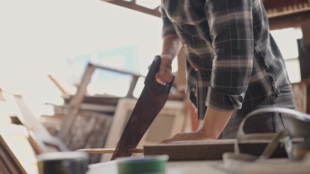 Skilled male carpenter wearing safety goggles using a hand saw to cut wood in a carpentry workshop. Focused craftsman working on timber furniture project in a rustic industrial studio