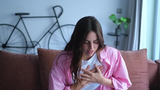 A young woman sits on a couch, experiencing noticeable chest pain and discomfort. She places her hands over her chest, then begins taking deep, calming breaths to find relief from the symptoms 
