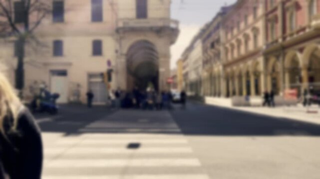 Defocused view of city life with a blonde woman waiting at a crosswalk while a red bus and scooters pass by on a sunny day, creating a dreamy and anonymous urban atmosphere