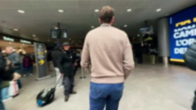 Back view of a young male tourist with a backpack walking through a modern airport terminal, passing by shops and other passengers on his way to the departure gate for his flight