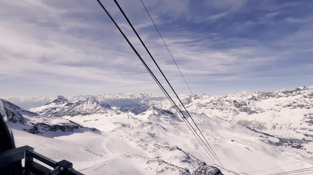 Personal perspective from a gondola moving up a mountain, offering a stunning panoramic view of the snow-covered alpine landscape and the valley below during a sunny winter day