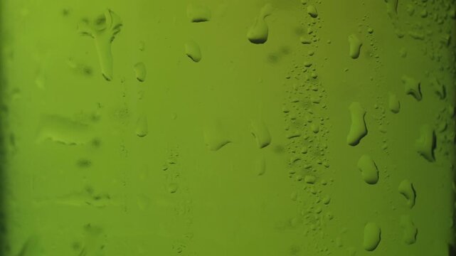Slow-motion macro shot of water droplets slowly trickling down the side of a cold, green glass bottle, showing condensation building up and drops merging for a fresh, cool abstract background