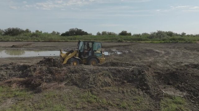 Wide view of small yellow excavator and backhoe loader working on large muddy construction site with deep tire tracks and green vegetation in background under blue sky