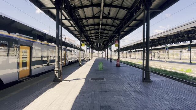 Sunny day view from a train departing an empty station, showing the long platform, railway tracks, and overhead canopy disappearing into the distance, conveying a sense of travel and solitude