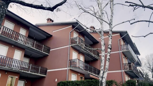 Smooth sliding shot revealing the facade of a modern residential building with balconies and windows. Seen through bare tree branches during a gray and overcast autumn day in a suburban neighborhood
