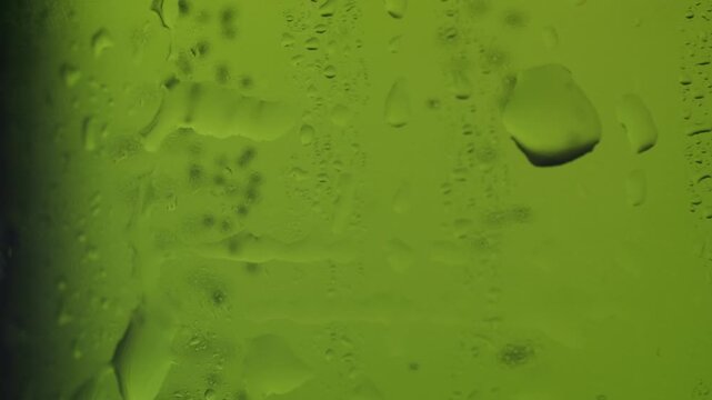 Slow-motion macro shot of water droplets slowly trickling down the side of a cold, green glass bottle, showing condensation building up and drops merging for a fresh, cool abstract background