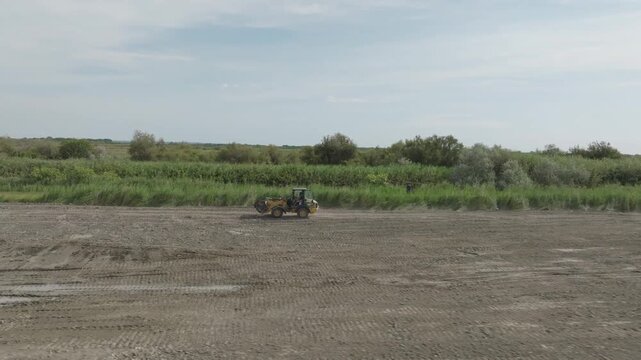 Yellow wheel loader driving on muddy dirt track in Camargue wetland with green vegetation in background under bright daylight